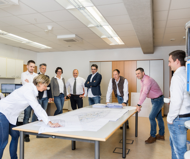 Neun Personen sitzen in einem modernen Büro um einen Tisch mit Bauplänen und besprechen ein Projekt.