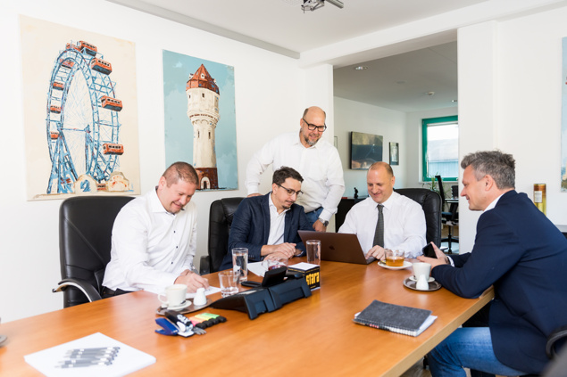 Fünf Männer in Geschäftskleidung treffen sich an einem Tisch mit einem Laptop, Dokumenten und Getränken.