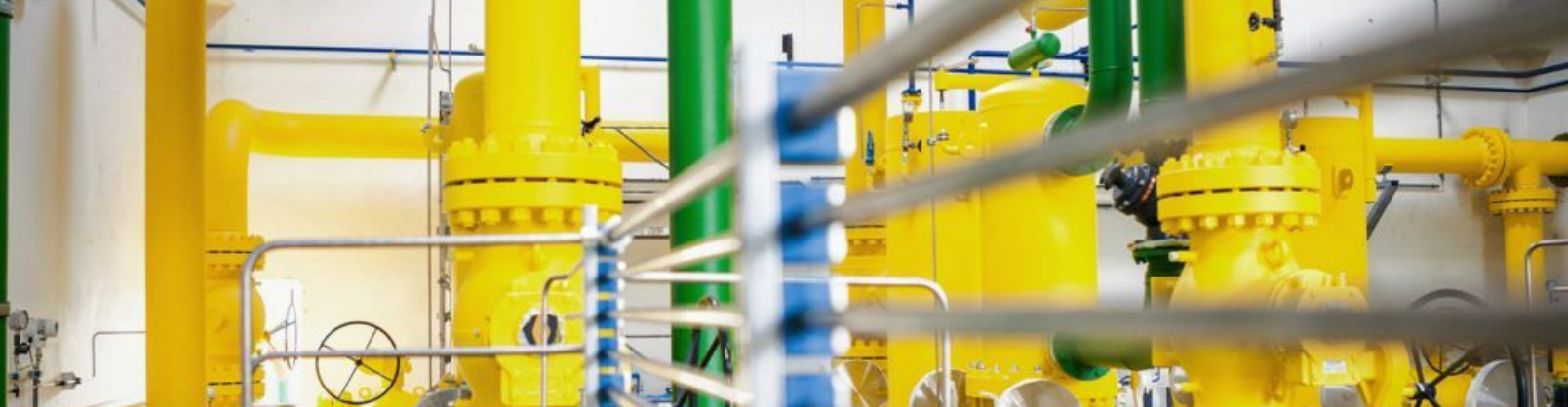 Gelbe und grüne Industrierohre und -ventile in einer hell erleuchteten Anlage, durch Metallgitter betrachtet.