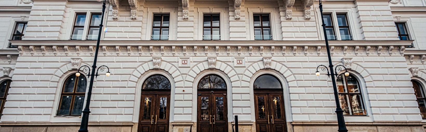 Verschnörkelte weiße Fassade mit drei Rundbogenportalen, hohen Fenstern und einem Treppenhaus mit schwarzem Geländer.