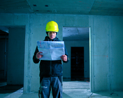 Person mit gelbem Schutzhelm studiert Baupläne in einem unfertigen Betongebäude.
