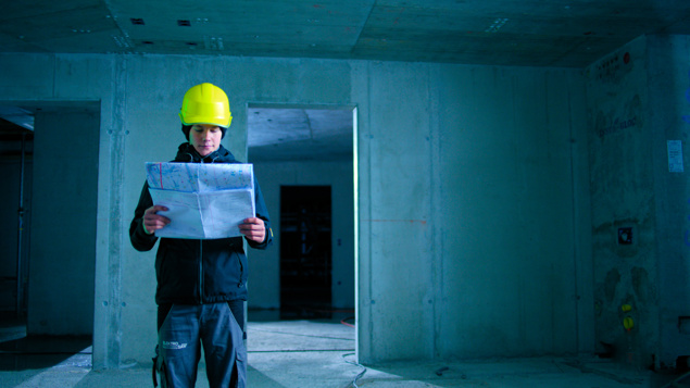 Person mit gelbem Schutzhelm studiert Baupläne in einem unfertigen Betongebäude.