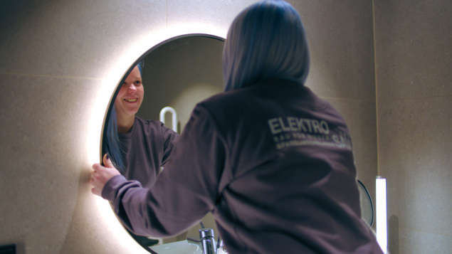 Eine Person mit blauen Haaren lächelt ihr Spiegelbild in einem runden, beleuchteten Spiegel in einem Badezimmer an.