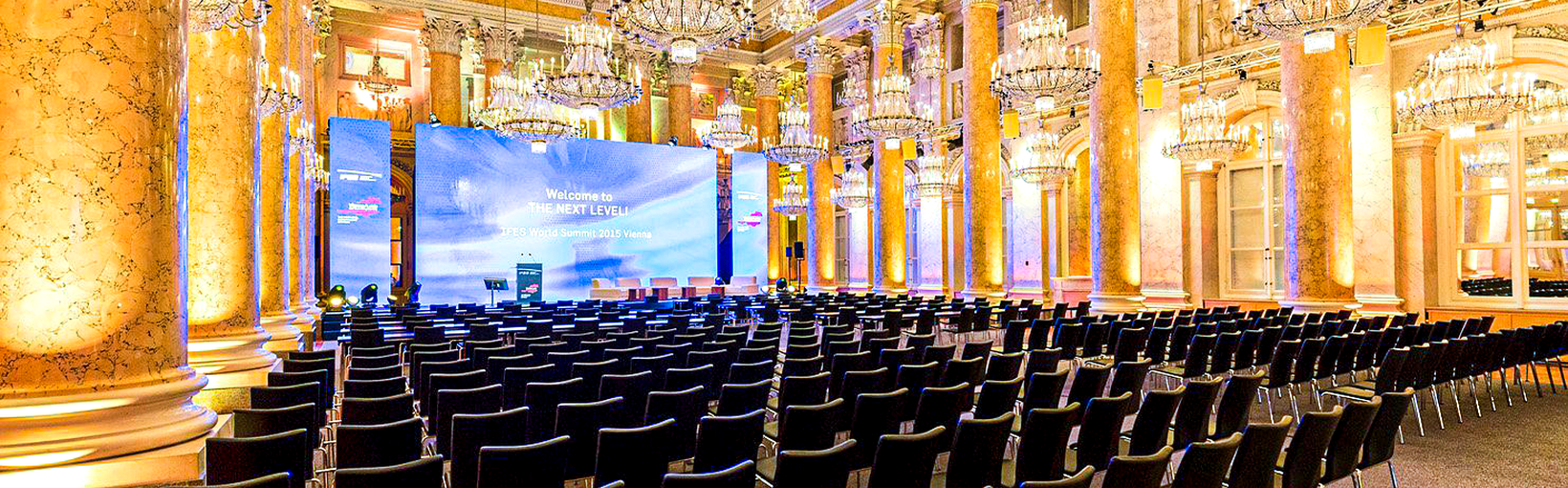 Großer Ballsaal mit leeren Stuhlreihen vor einer großen Bühne, verschnörkelten Säulen und eleganten Kronleuchtern.