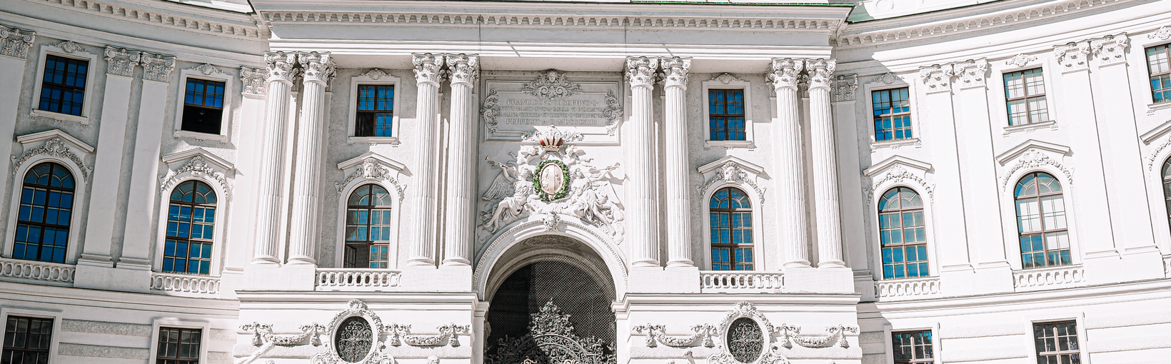 Barockes Gebäude mit grüner Kuppel, Pferdekutschen und Menschen davor unter blauem Himmel.
