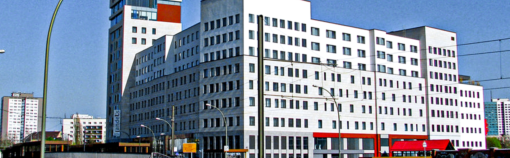 Großes weißes Bürogebäude mit vielen Fenstern neben einer Straße und Bahngleisen an einem klaren, sonnigen Tag.