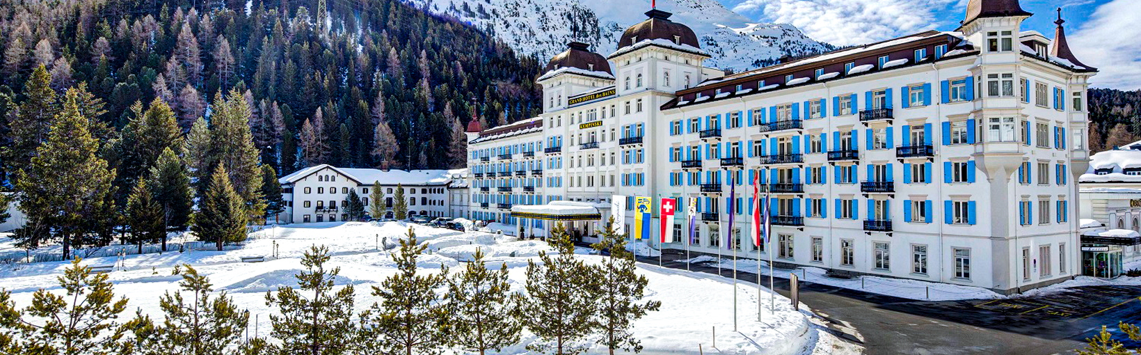 Grand Hotel mit blauen Fensterläden in verschneiter Berglandschaft unter strahlendem Himmel, umgeben von Kiefern.