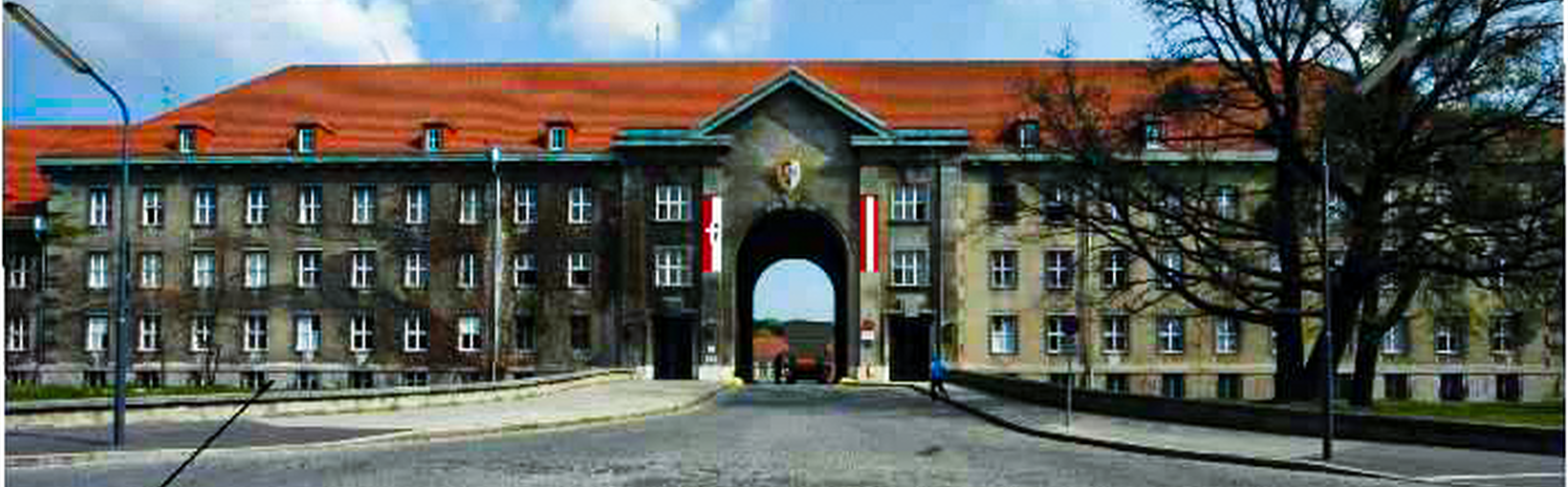 Großes Backsteingebäude mit zentralem Torbogen, rotem Dach und zwei vertikalen rot-weißen Bannern auf beiden Seiten.