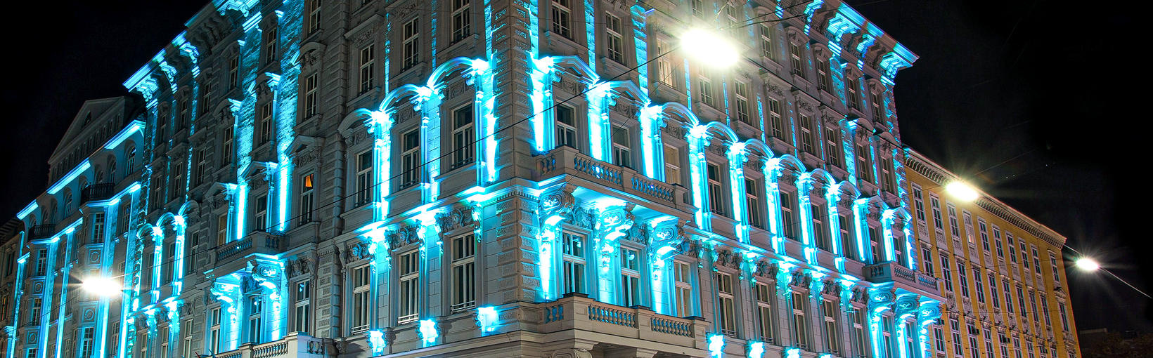 Ein großes verziertes Gebäude, das nachts mit blauen Lichtern beleuchtet wird, mit Lichtspuren von vorbeifahrenden Autos.