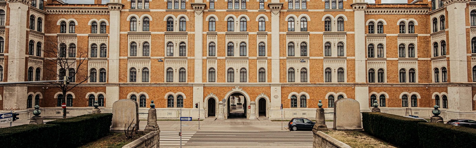 Ein großes historisches Backsteingebäude mit Rundbogenfenstern und Türmen, das man von einem Weg aus sieht, der zum Eingang führt.
