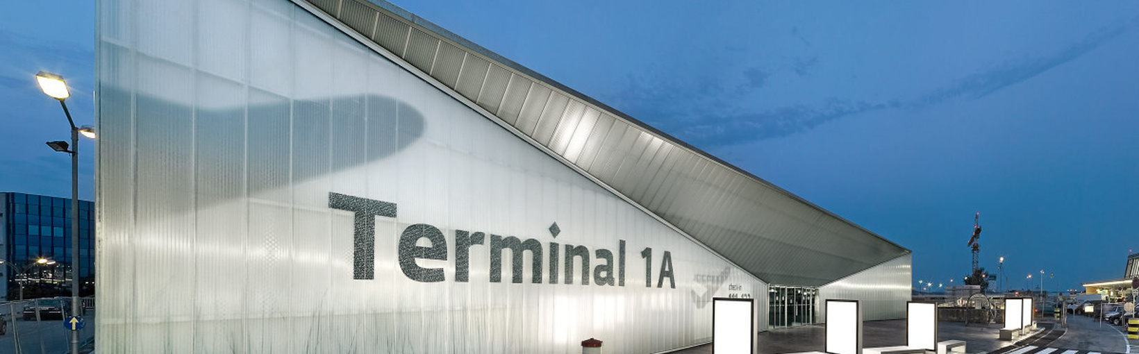 Modernes Glasgebäude mit Terminal 1A-Schild auf einem Flughafen, abends beleuchtet.