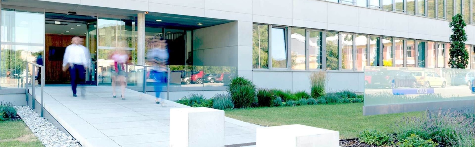 Menschen betreten ein modernes Bürogebäude mit Glastüren und Sitzwürfeln im Freien auf einem gepflasterten Weg.