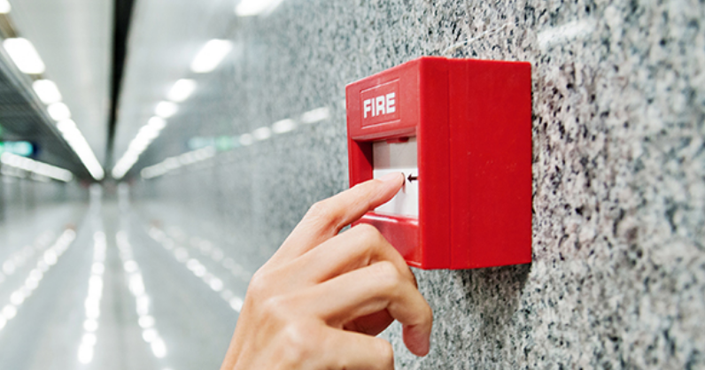 Eine Hand drückt auf einen roten Feuermelder an einer Marmorwand in einem Flur.