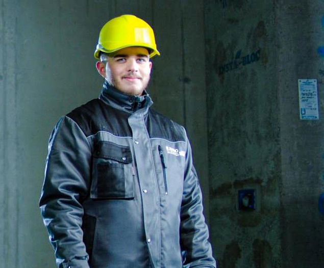 Ein Bauarbeiter mit einem gelben Schutzhelm und einer dunklen Jacke steht in einem Gebäude, schaut in die Kamera und lächelt.